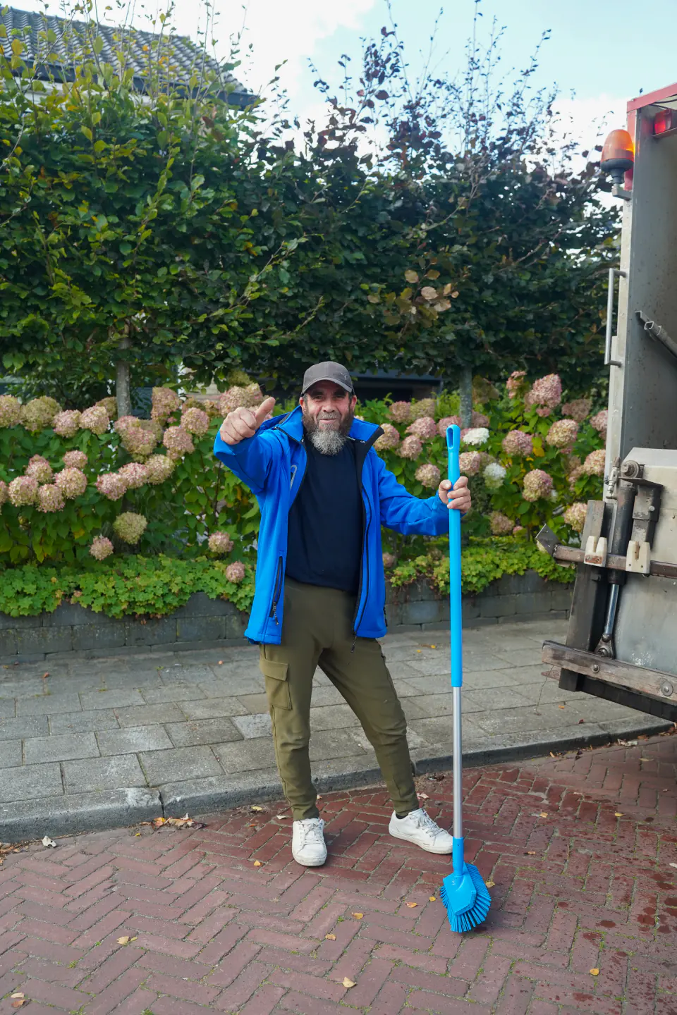 Medewerker van Frisse Kliko met pet en blauwe jas naast de reinigingswagen.